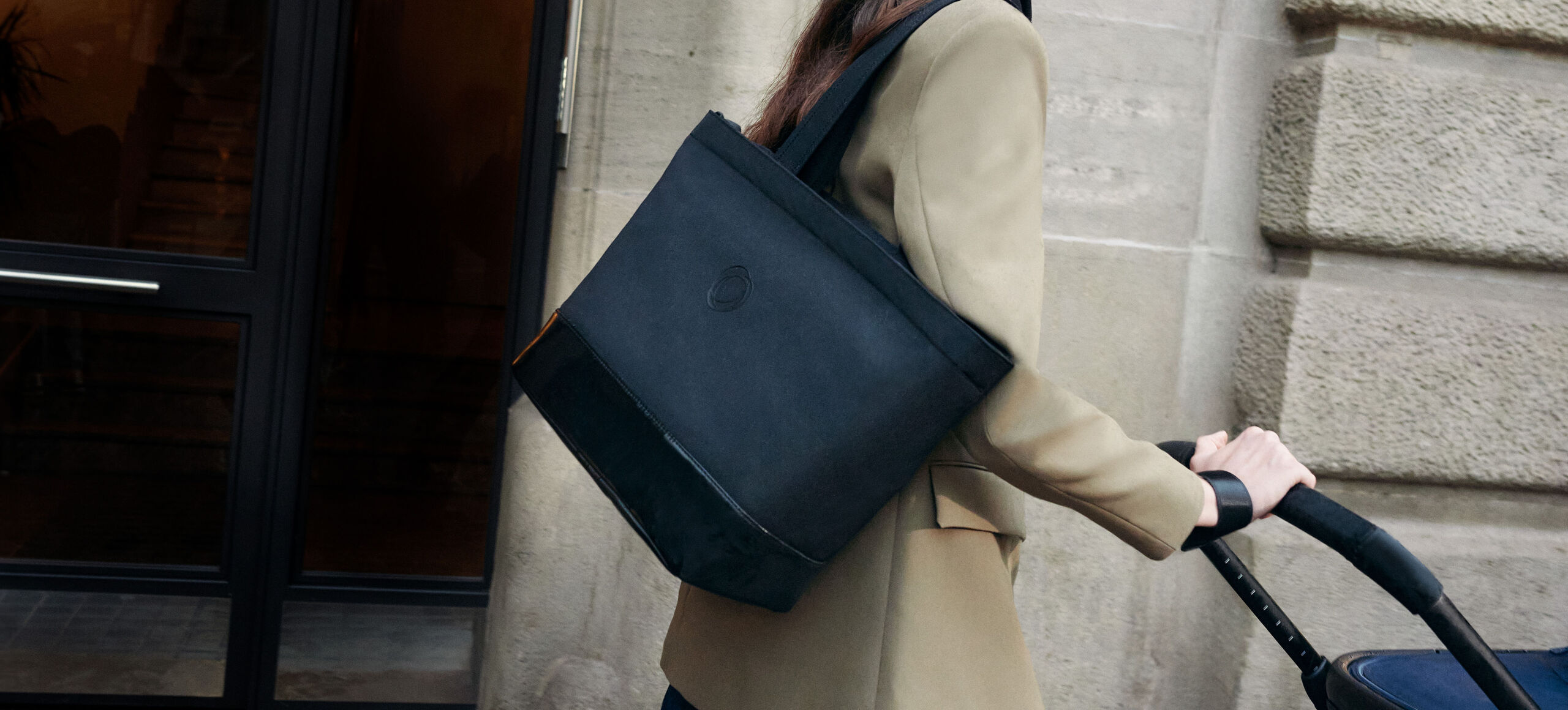 Eine Frau geht an einem Steingebäude entlang und schiebt einen Bugaboo Kinderwagen, während sie eine große schwarze Bugaboo Wickeltasche über ihrer Schulter trägt.