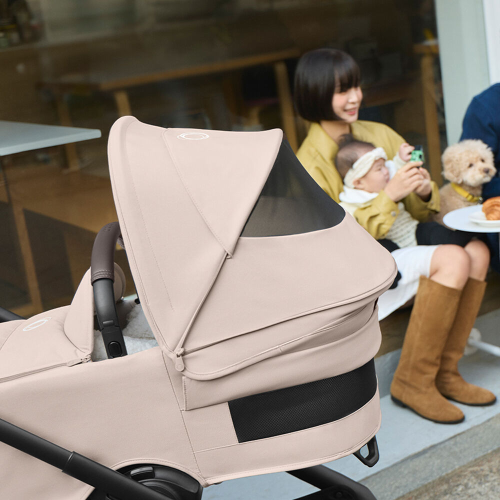 A Bugaboo Dragonfly Plus city stroller in Desert Taupe in the foreground, with a mom holding her baby seated in the background.