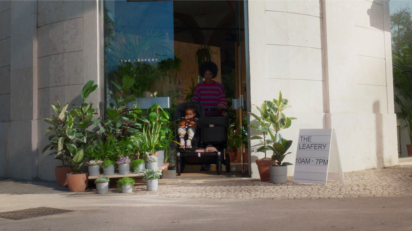 Een moeder en haar kinderen stappen uit een café omringd door weelderige groene planten bij de ingang. De kinderen zitten in de Bugaboo Donkey 5 Duo, met één kind in een stoel en de andere in de wieg.