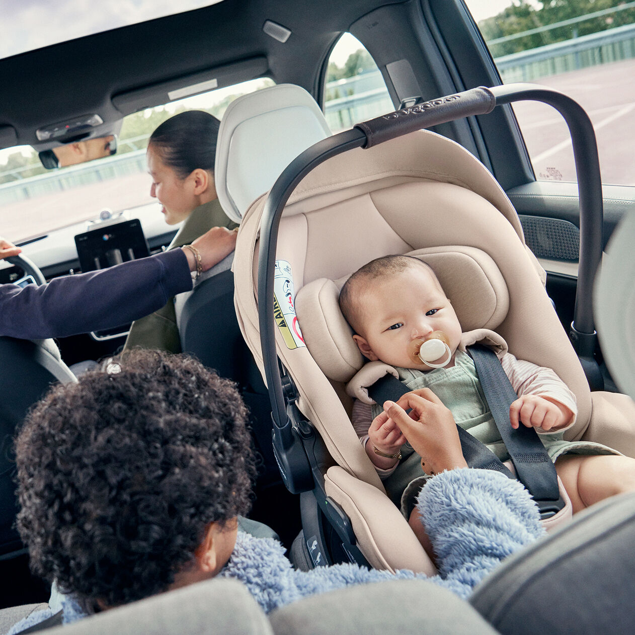 Un bébé suçant sa tétine est installé en toute sécurité dos à la route dans le siège auto pour nouveau-né Bugaboo Otter by Nuna. Son frère est assis à côté d'elle et lui tient la main. Leurs parents sont installés à l'avant de la voiture.
