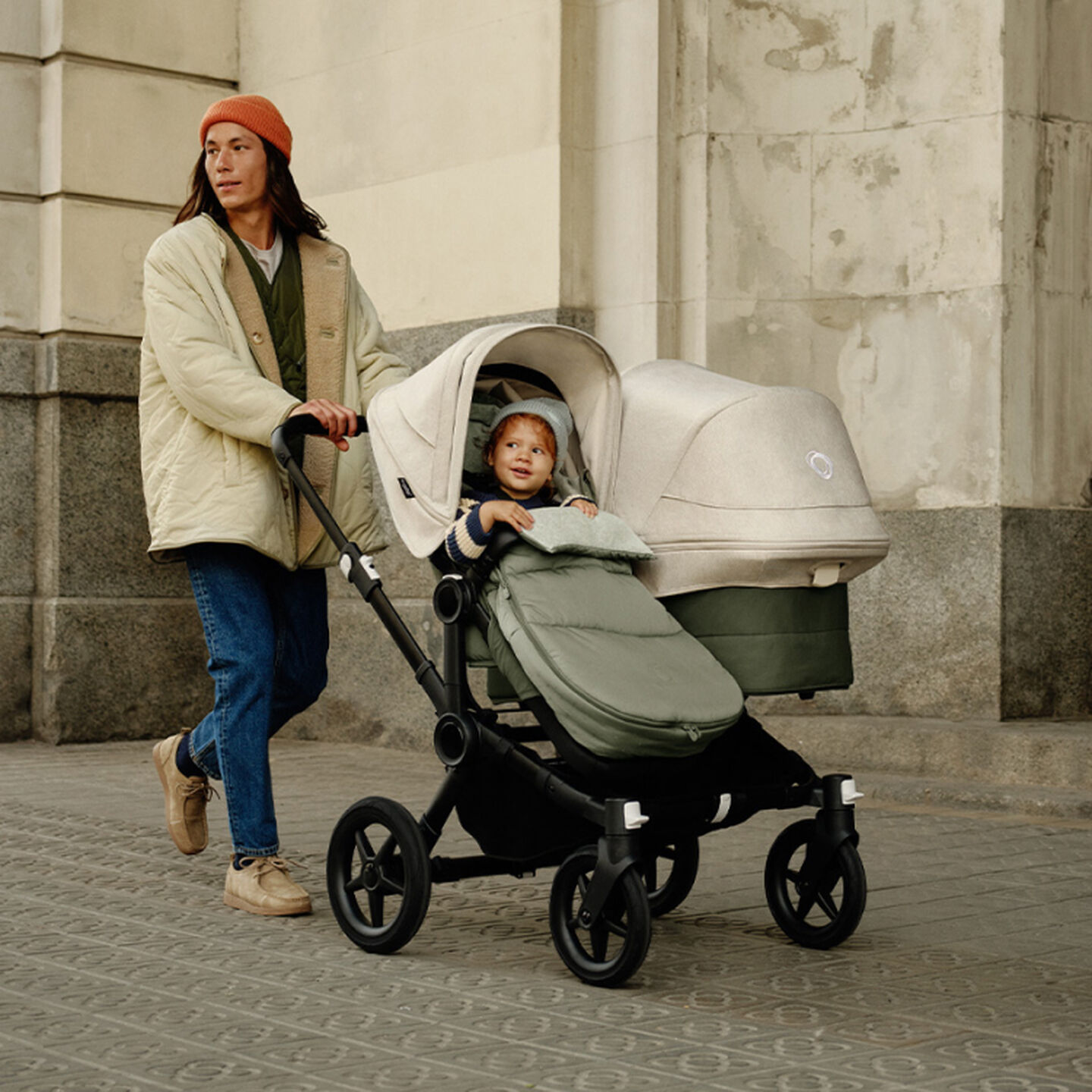 A father walks with his kids in the Bugaboo Donkey 5 Duo stroller. His toddler sits on the left seat inside a footmuff. The bassinet on the right faces the other way, showing the Bugaboo logo on the canopy.
