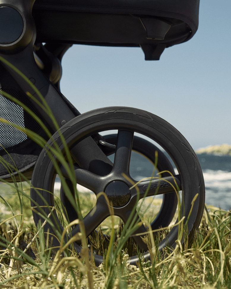 Close-up of the all-terrain wheels of a Bugaboo stroller as it moves across a grassy area, with a scenic coastline and sea in the background.
