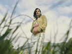 A pregnant mom stands in a grassy field with one hand on her bare baby bump, with mountains in the background and tall blades of grass in the foreground.
