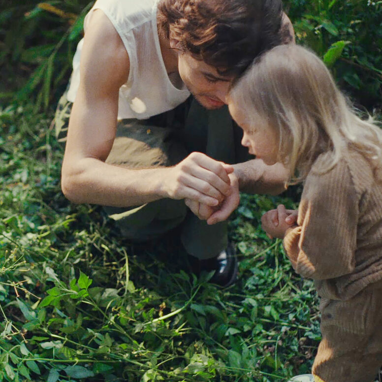 Un père et sa fille sont accroupis dans un jardin sauvage envahi de végétation. Il lui montre ce qu'il a dans ses mains.