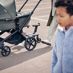 A Bugaboo Fox 5 Renew stroller on the street with a comfort wheeled board attached. In the foreground, a child walks by, appearing blurred.