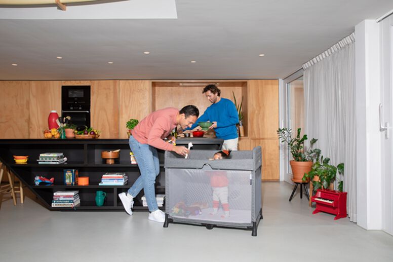 Two men are in the kitchen - one is cooking and the other is playing with a toddler, who is standing in the Bugaboo Stardust travel cot.