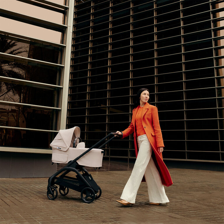 A stylish mom stands next to a Bugaboo Dragonfly city stroller with a bassinet and fabrics in Desert Taupe. Behind her is a tall, modern building accentuated with horizontal lines.