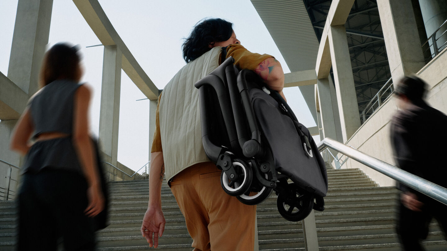 Ein Vater trägt einen zusammengeklappten Bugaboo Kompaktkinderwagen auf einer Schulter, während er auf die Treppe zugeht.