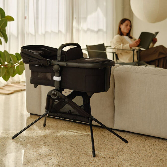 A Bugaboo bassinet placed on a bassinet stand, inside a cozy living room. In the background, a mom is reading and relaxing.