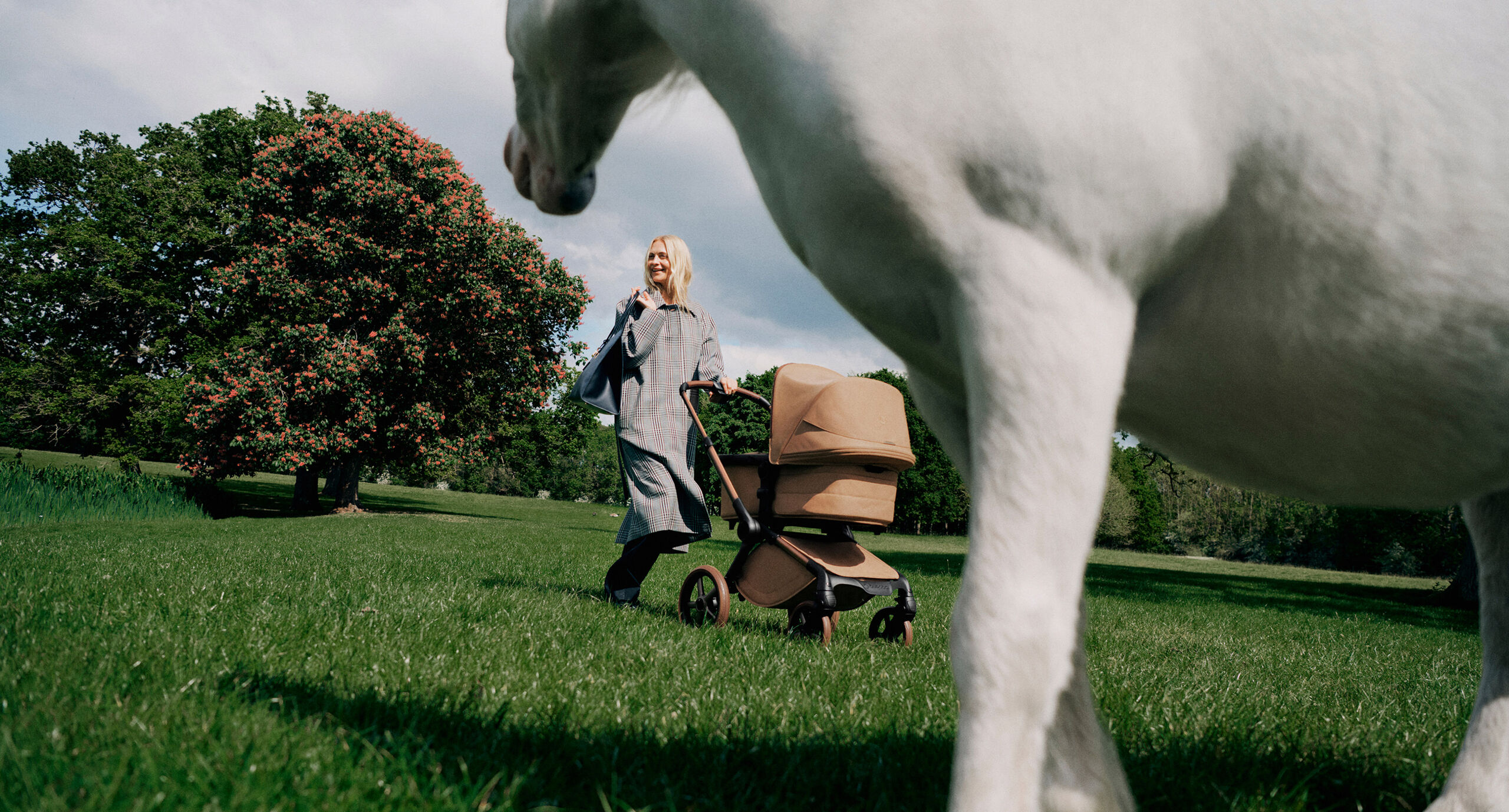 Une maman élégante pousse une poussette Bugaboo x Stella McCartney dans un parc avec des arbres verdoyants et un ciel bleu en arrière-plan. Au premier plan, partiellement hors cadre et flou, se trouve un poney blanc.