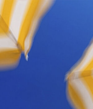 A blurred close-up of yellow and white striped beach umbrellas against a bright blue sky.