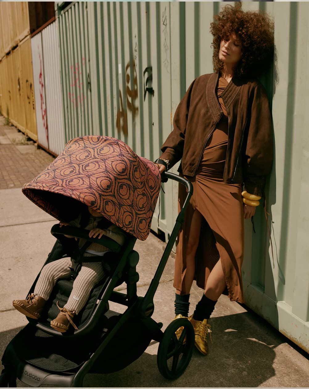 A stylish mom stands outdoors, leaning casually against the side of a metal container. Next to her, her child sits inside a Bugaboo Fox 5 Renew stroller with a Bugaboo X Artipoppe Limited Edition sun canopy.
