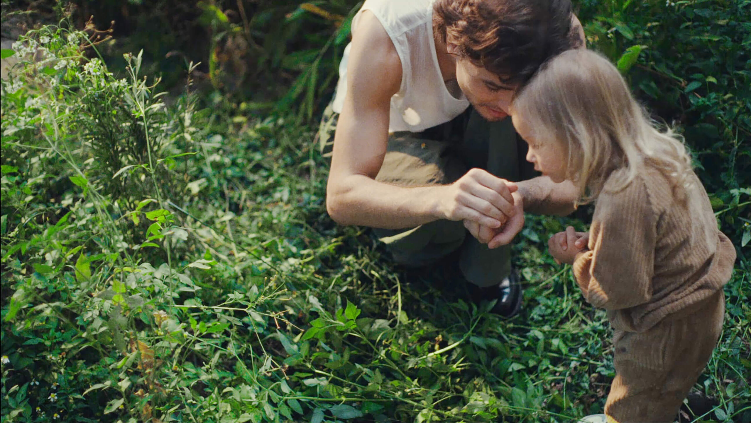 A dad and his daughter crouch in an untamed, overgrown garden. He's showing her something he's caught in his hands.