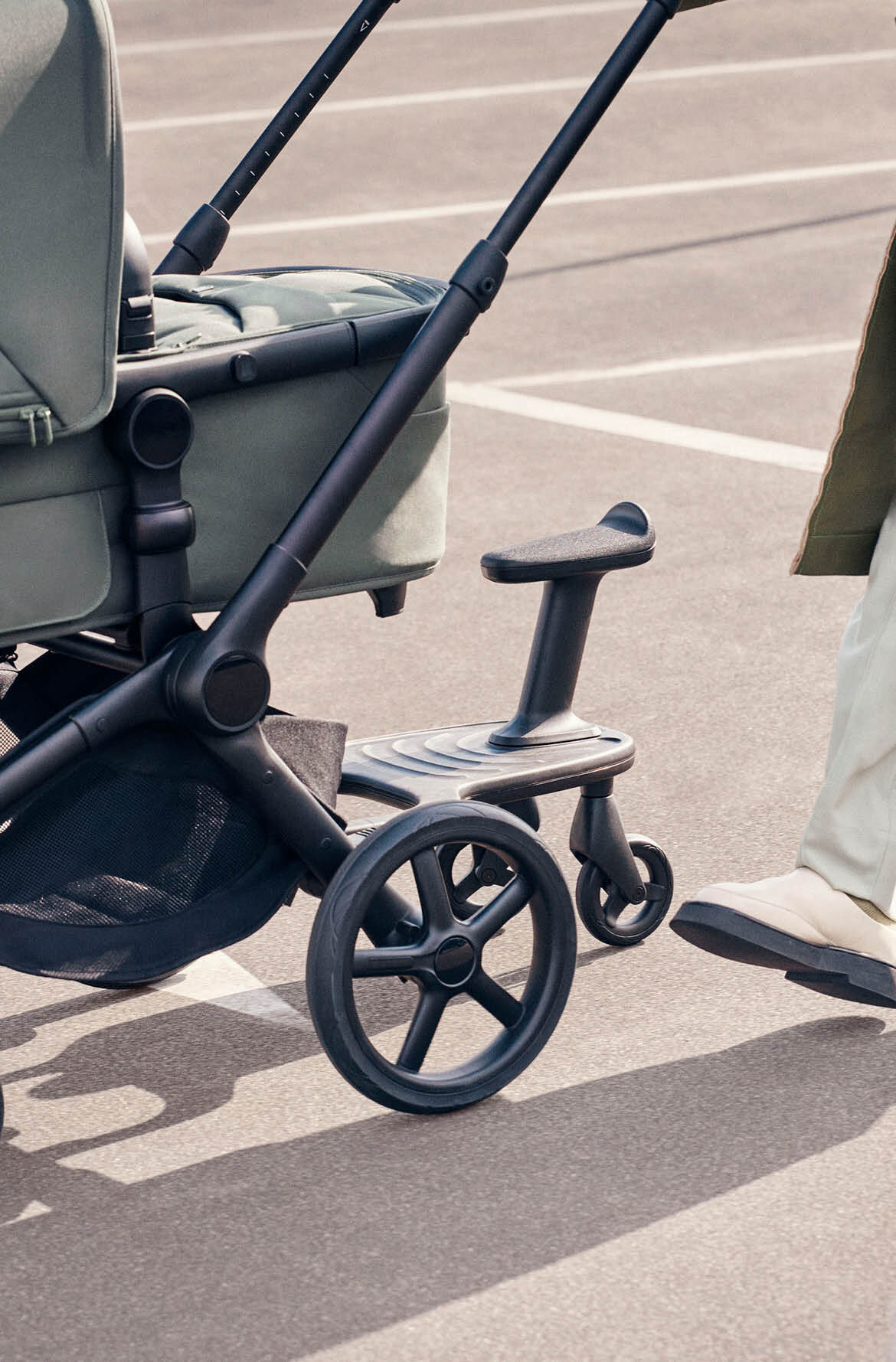 A parent pushes a Bugaboo Fox 5 Renew bassinet stroller with a Bugaboo wheeled board attached, while a child walks alongside.