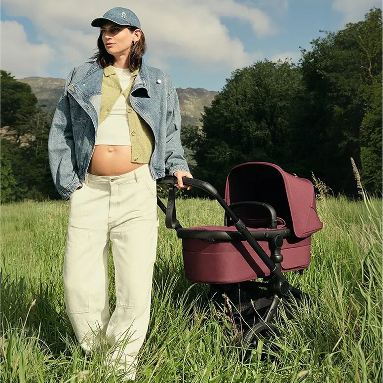 A stylish mom with a pregnant belly stands in a grassy field next to a Bugaboo Fox 5 Renew stroller with Dark Cherry fabrics. A mountain range sits in the distance.
