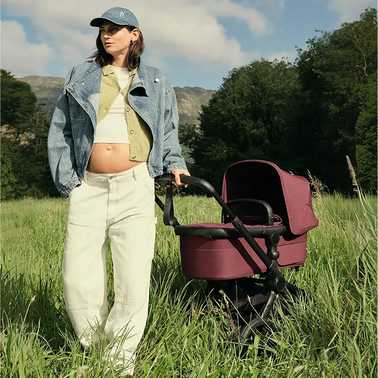 A stylish mom stands next to a Bugaboo Dragonfly city stroller with a bassinet and fabrics in Desert Taupe. Behind her is a tall, modern building accentuated with horizontal lines.