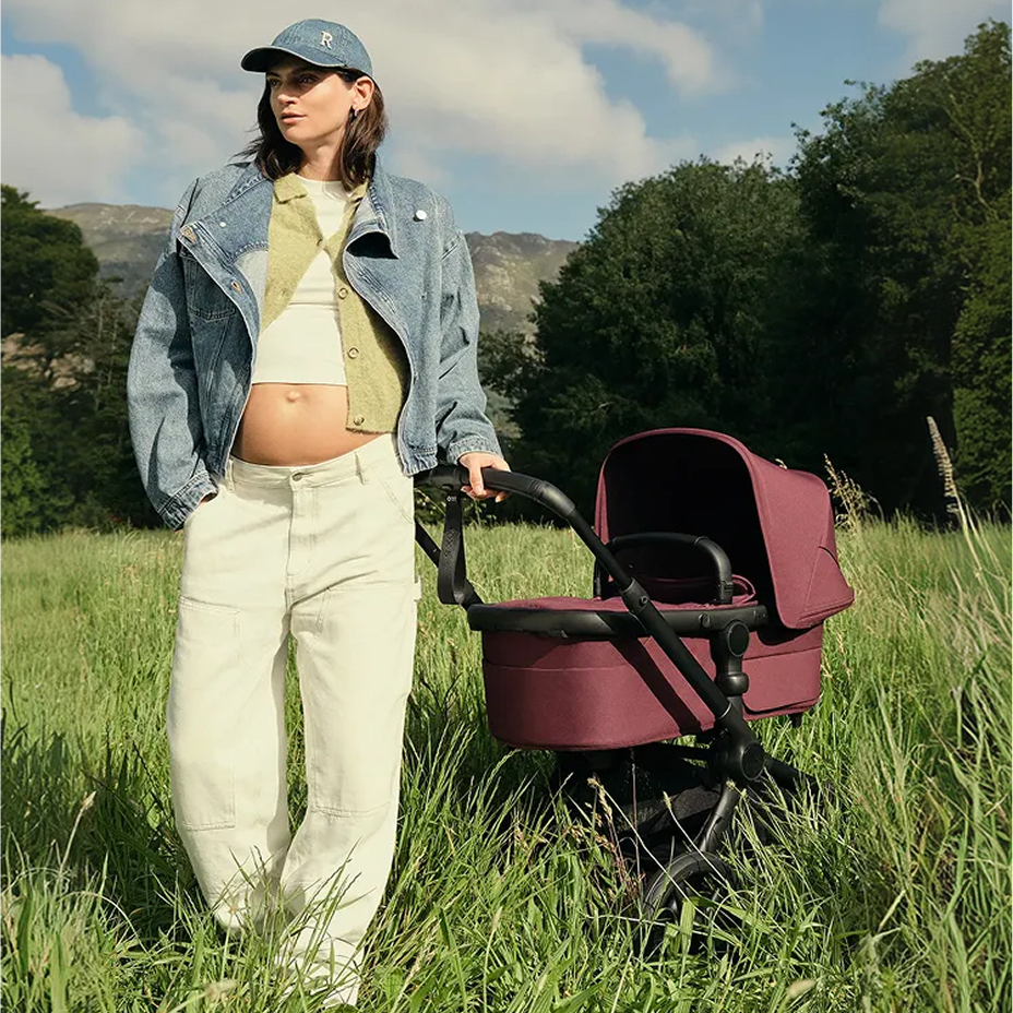 A stylish mom stands next to a Bugaboo Dragonfly city stroller with a bassinet and fabrics in Desert Taupe. Behind her is a tall, modern building accentuated with horizontal lines.