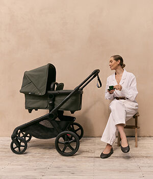 A mom drinking tea sits beside a green Bugaboo Kangaroo bassinet stroller, against a minimalist outdoor backdrop.