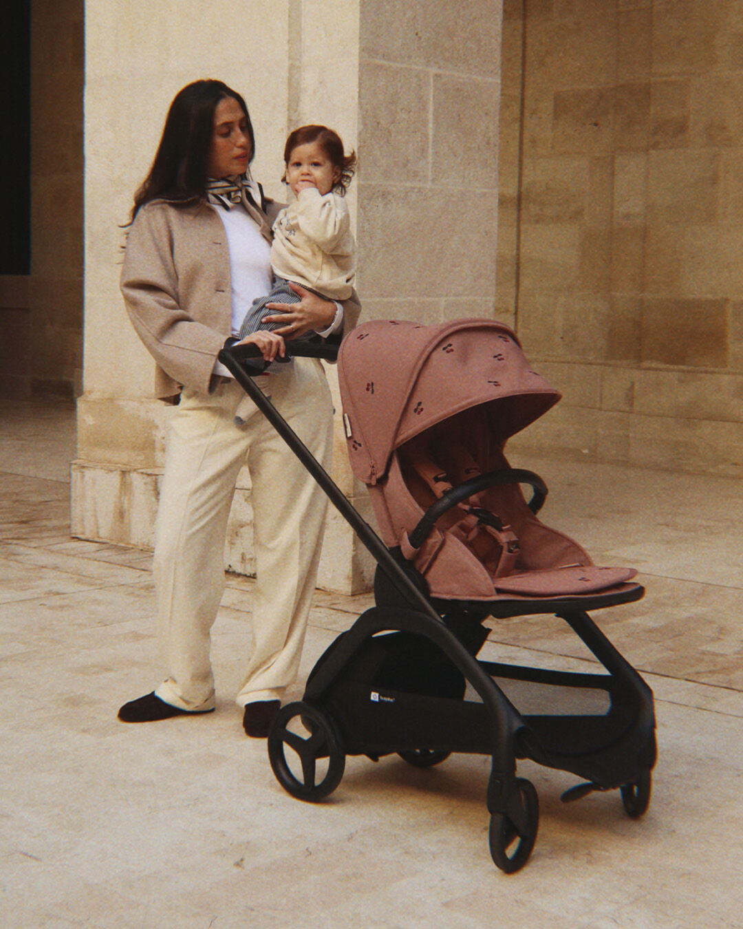 A smiling mom standsvwith her bab next to a Bugaboo Dragonfly pushchair. The stroller features limited-edition fabric embroidered with Konges Sløjd cherry motif. They’re in a green courtyard outside a stone-walled building.