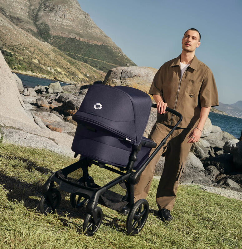 A mom pushes a Bugaboo Donkey 6 that carries three children: One on a seat, one in the bassinet, and a third child riding along with an attached wheeled board.