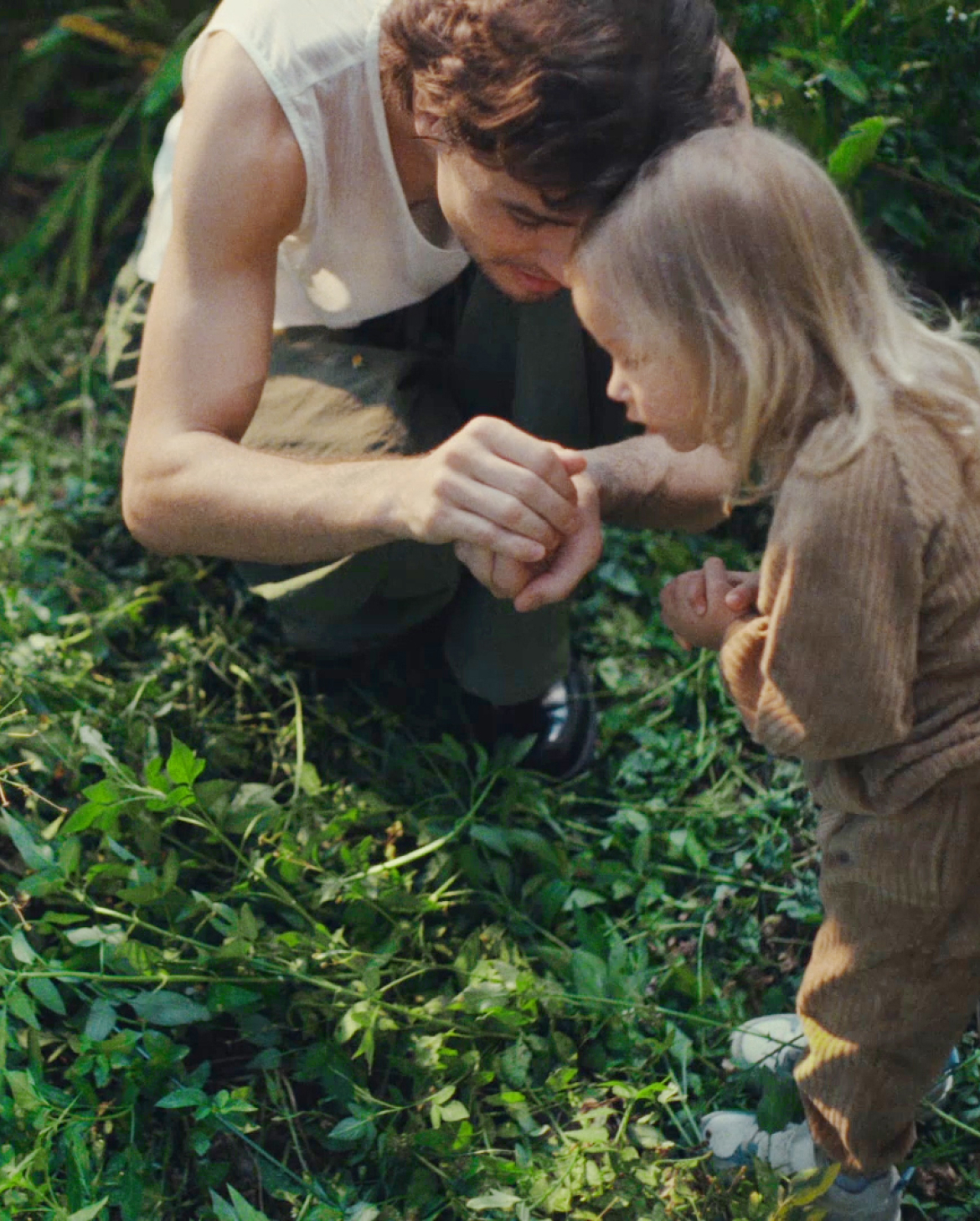 A dad and his daughter crouch in an untamed, overgrown garden. He's showing her something he's caught in his hands.