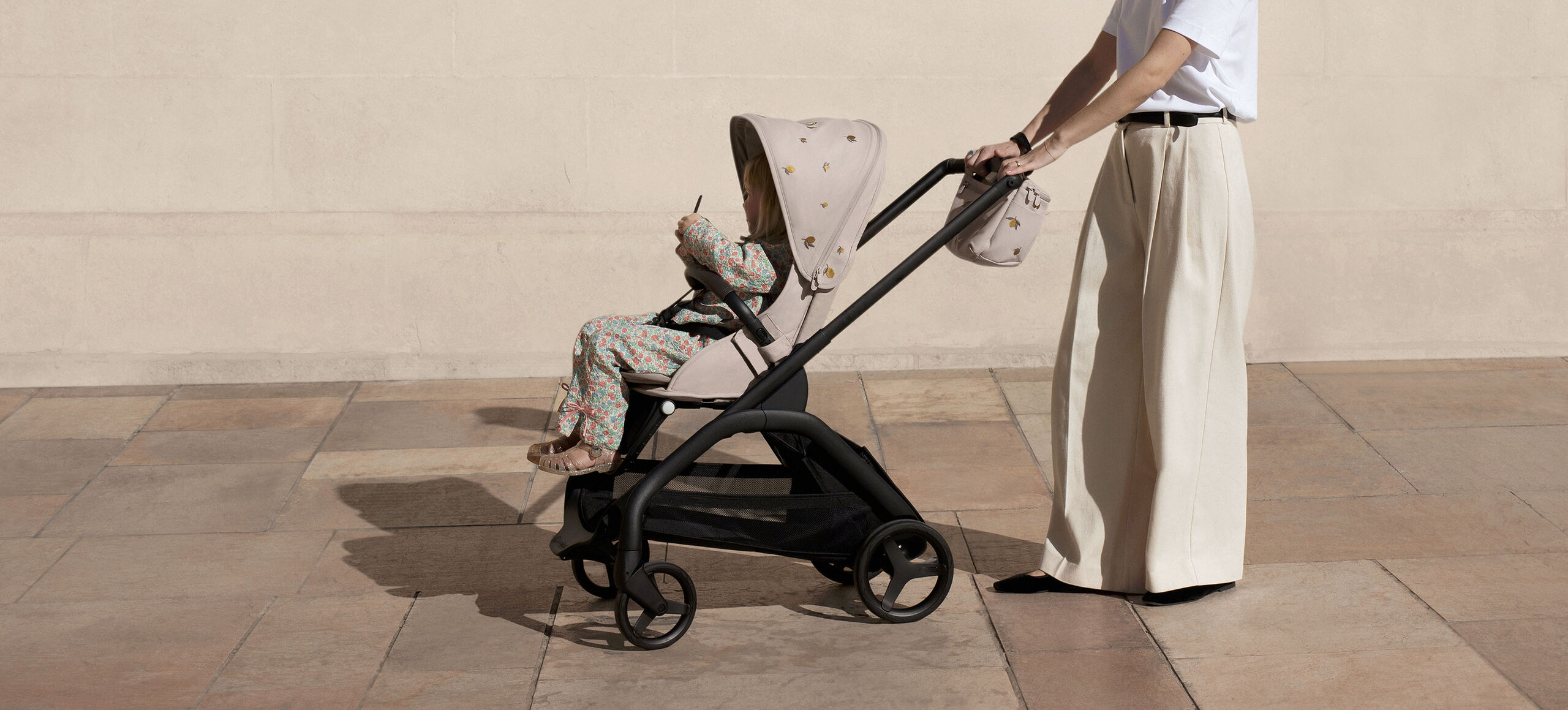 A mother strolls with her toddler sitting in a Bugaboo Dragonfly seat stroller. The stroller features a Bugaboo X Konges Sløjd Limited Edition sun canopy and organizer.