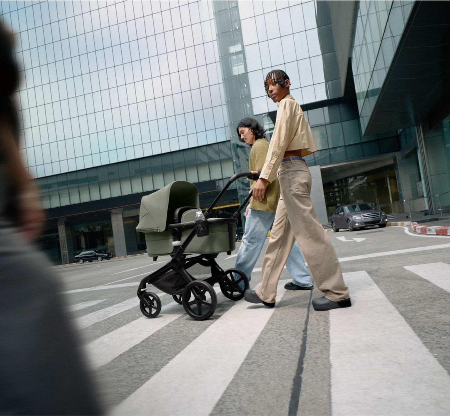 スタイリッシュなカップルが、バガブー フォックス 5 ストローラーに新生児を乗せて自信に満ちた足取りで歩いている。 周りにはガラス張りの高層ビルが建ち並んでいる。