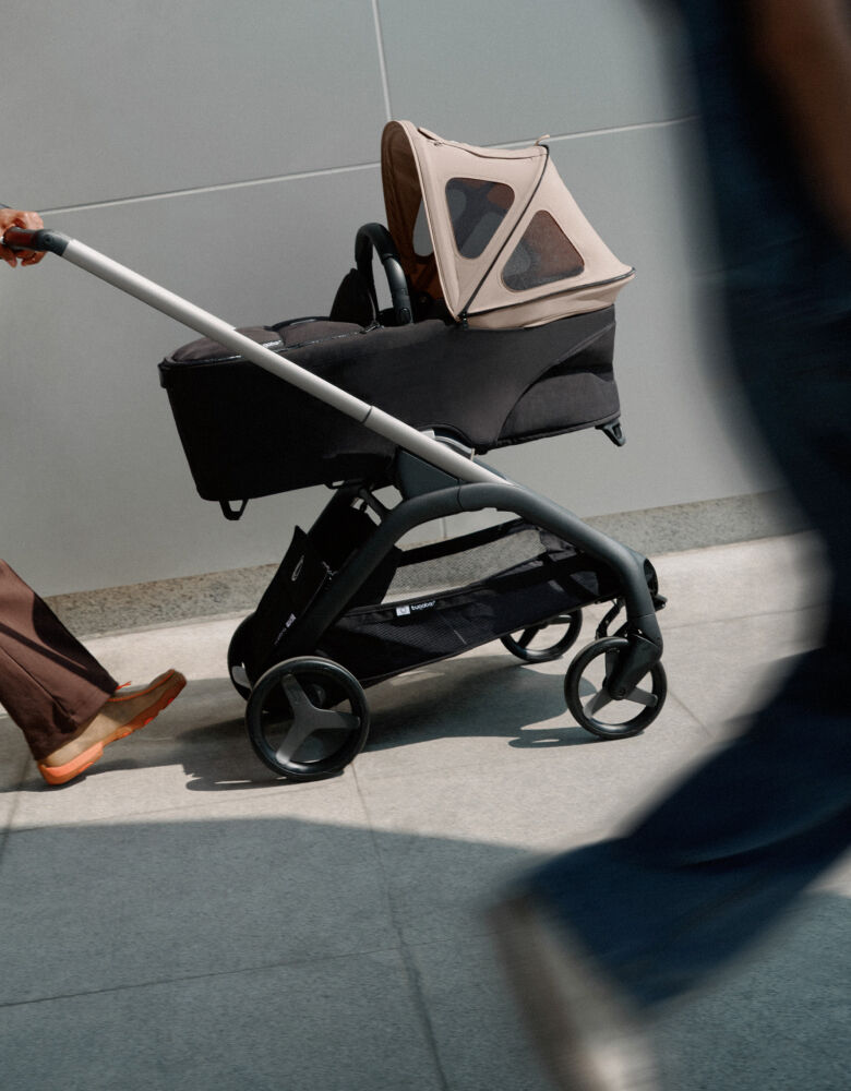Une poussette Bugaboo avec une nacelle et une capote à fenêtres sur le trottoir. L'image est légèrement inclinée, avec des ombres floues en premier plan.