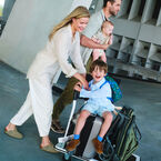 Alt A happy family on their way for a trip. Dad carries the baby in his arms while Mom pushes a luggage trolley with the toddler sitting on top of a suitcase. The trolley is also stacked with the Bugaboo Butterfly 2 stroller.