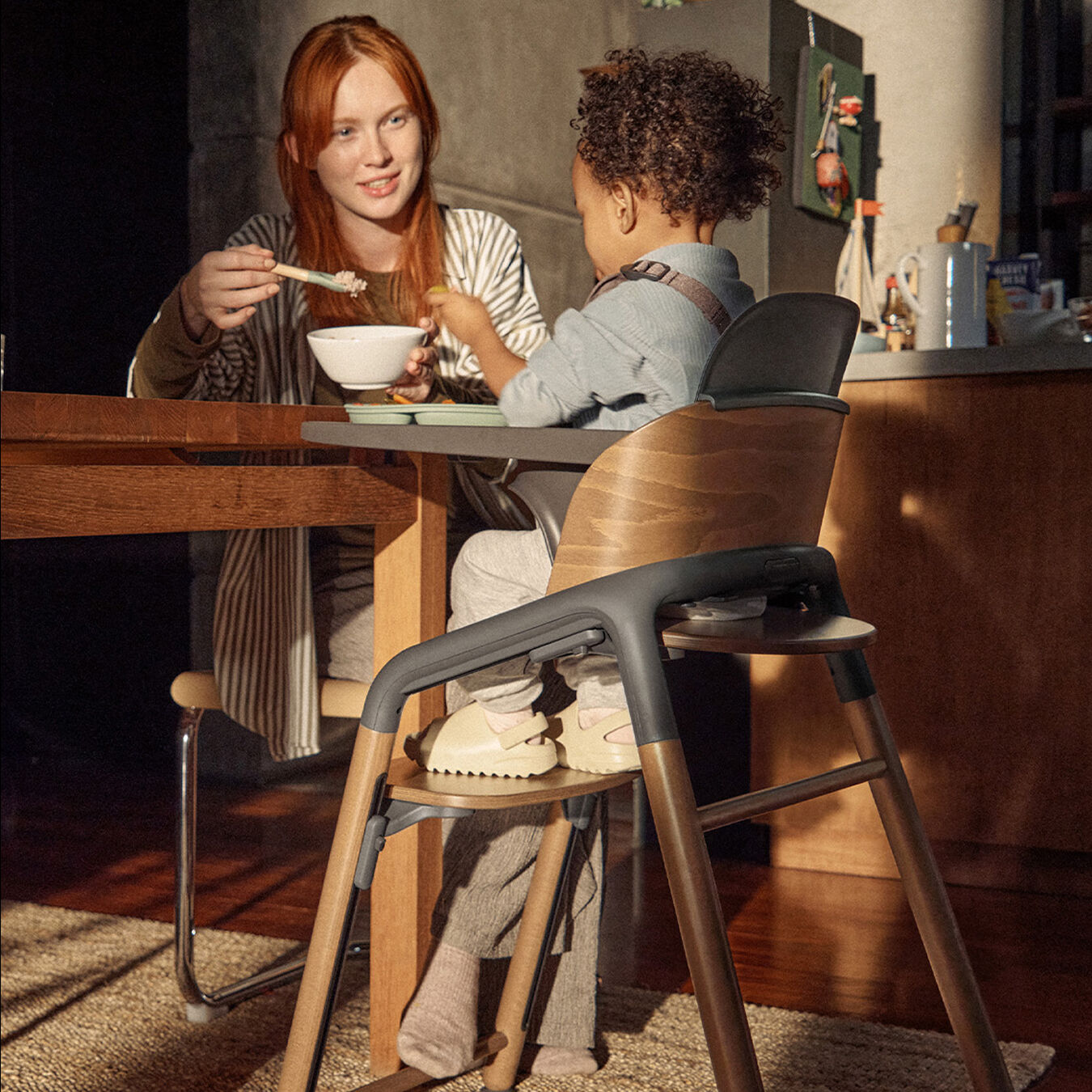 A mom feeds her toddler who’s seated in a Bugaboo Giraffe high chair with a tray attached. They’re sitting in a warm, softly lit kitchen with wooden furniture.