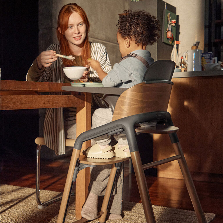 A mom feeds her toddler who’s seated in a Bugaboo Giraffe high chair with a tray attached. They’re sitting in a warm, softly lit kitchen with wooden furniture.