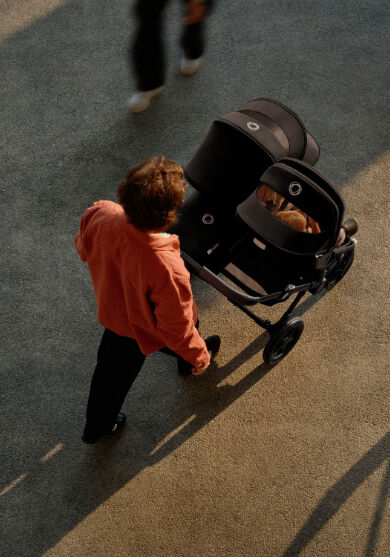 A top-down view of a father strolling with his two children in a Bugaboo Donkey 5 double stroller, with a seat and a bassinet in black fabrics. A toddler is visible through the peek-a-boo panel of the seat's sun canopy.