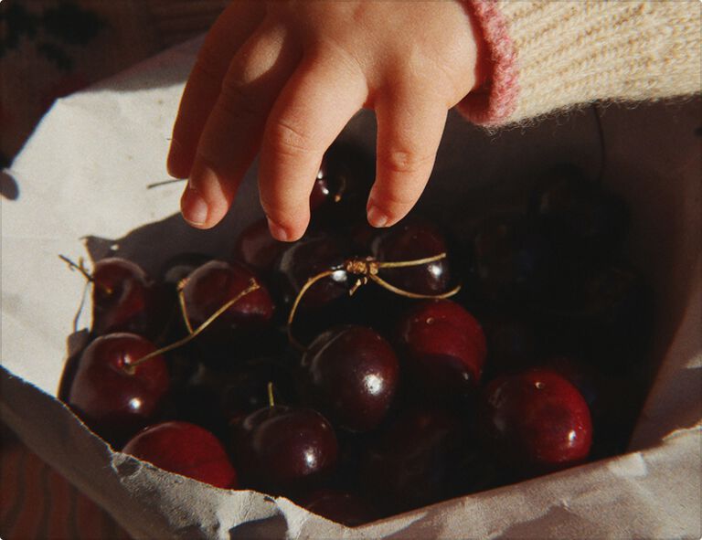 A tiny baby’s hand reaches into a bowl full of red cherries.