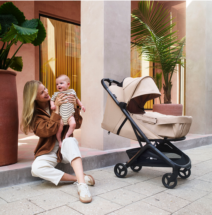 Une maman soulève son bébé devant une véranda bordée d'arbres en pot. À côté d'eux se trouve la poussette Bugaboo Butterfly 2 en tissu Desert Taupe, équipée d'un cocon pour nouveau-nés.