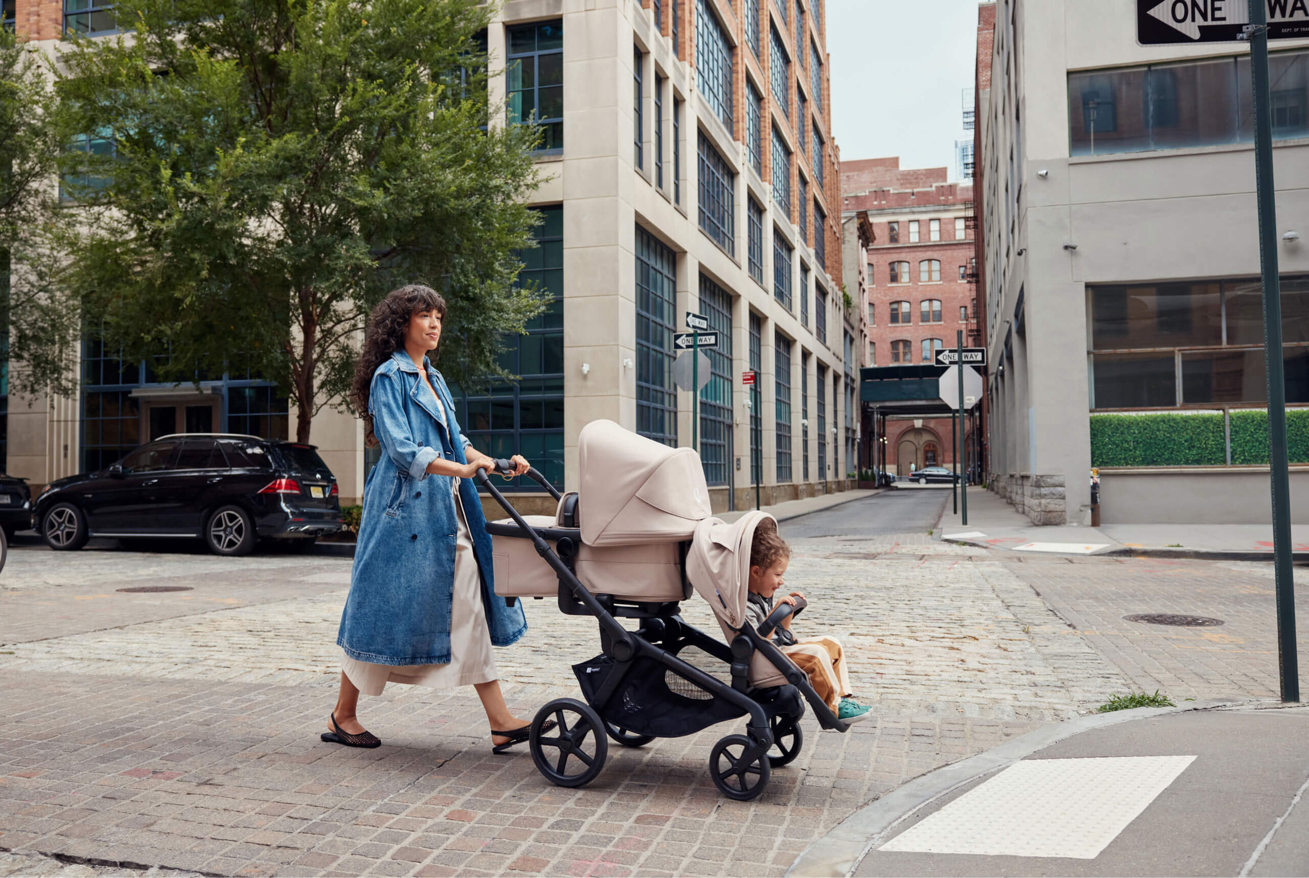 A mother walks down the street, pushing a Bugaboo Kangaroo tandem double pram with a bassinet facing parent and a seat facing the world. A toddler is sitting in the seat. They're in an urban setting with buildings in the background.