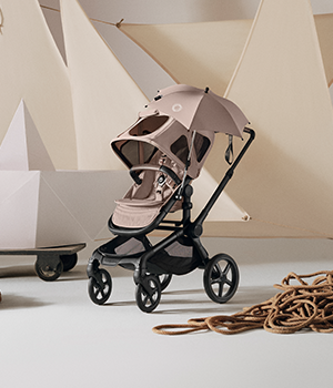 A Bugaboo Fox 5 Renew seat stroller in Desert Taupe fabric with a matching breezy sun canopy and parasol stands against a backdrop of cream fabric panels. On the ground are piles of ropes and stage props.