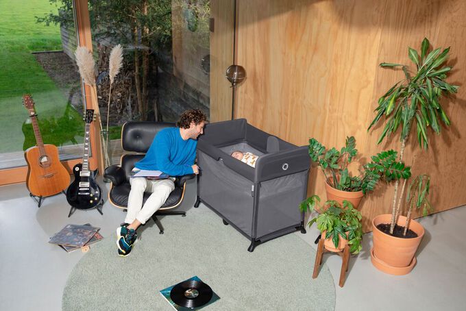 : A man sitting in a swivel chair in his music studio, looking over at a baby lying in the bassinet insert of the Bugaboo Stardust travel cot.
