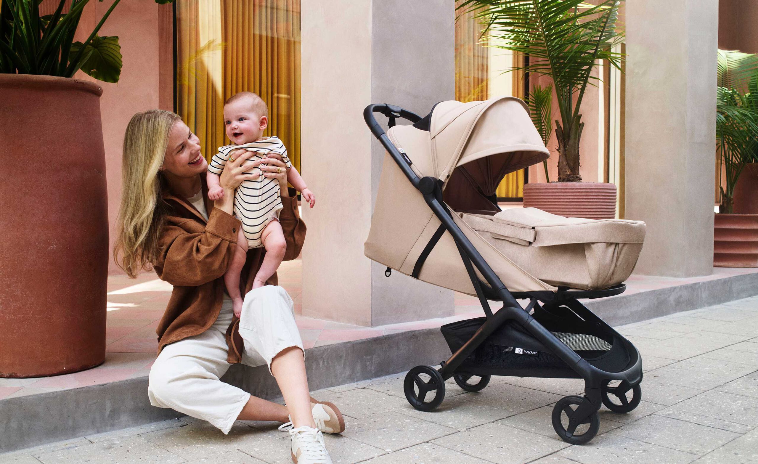 Une maman soulève son bébé devant une véranda bordée d'arbres en pot. À côté d'eux se trouve la poussette Bugaboo Butterfly 2 en tissu Desert Taupe, équipée d'un cocon pour nouveau-nés.