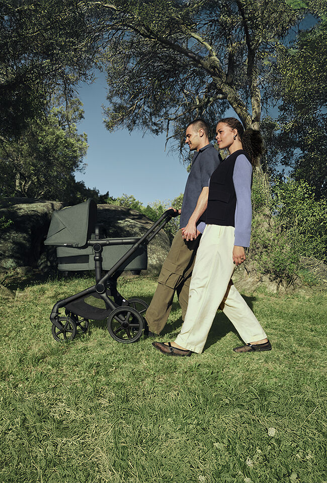 A family enjoys their day out at the park. The parents walk side by side, pushing a Bugaboo Kangaroo Fox 5 Renew bassinet stroller with Forest Green fabrics.