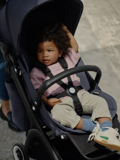 A toddler relaxes in the seat of an ultra-compact Bugaboo Butterfly travel pushchair. The toddler is securely seated with the harness fastened and a Bugaboo Butterfly bumper bar across their middle. 