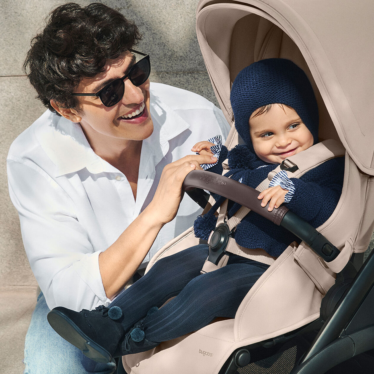 A smiling dad and baby on a sunny day. The baby wears a blue onesie and sits in a Bugaboo Dragonfly Plus stroller with Desert Taupe fabrics.