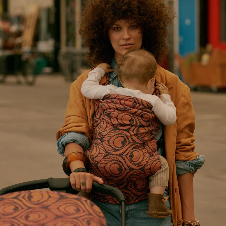 A stylish mom carries her baby in an Artipoppe Zeitgeist Baby Carrier with fabric that matches the Bugaboo stroller in front of them. The baby rests against her chest while she looks ahead calmly. Behind them is a casual, urban setting.