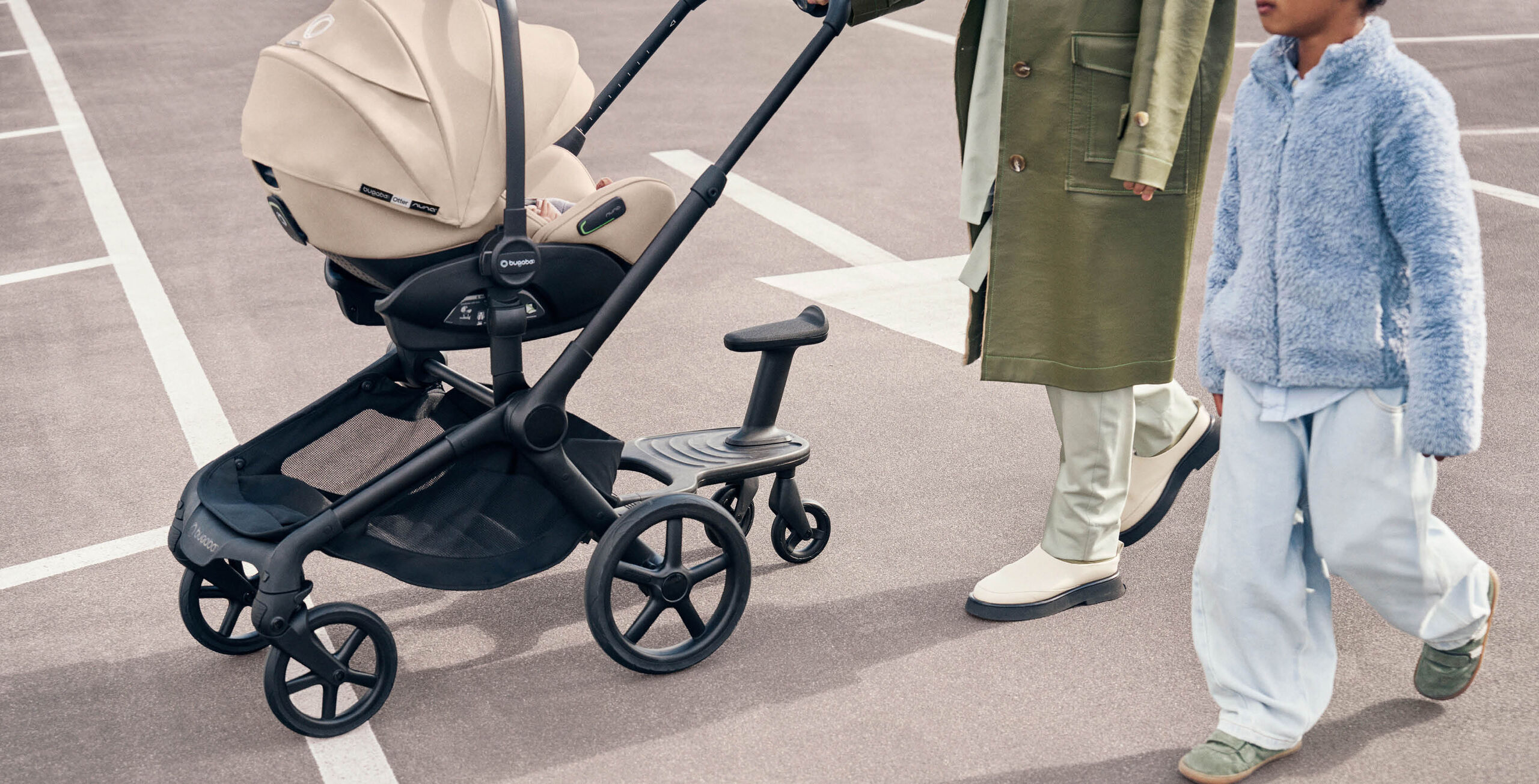 A parent pushes a Bugaboo pushchair with a Bugaboo Otter car seat and a Bugaboo wheeled board attached, while a child walks alongside.