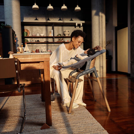 A casually dressed mom plays with her baby in a Bugaboo Giraffe high chair. They're in a warmly lit modern kitchen.