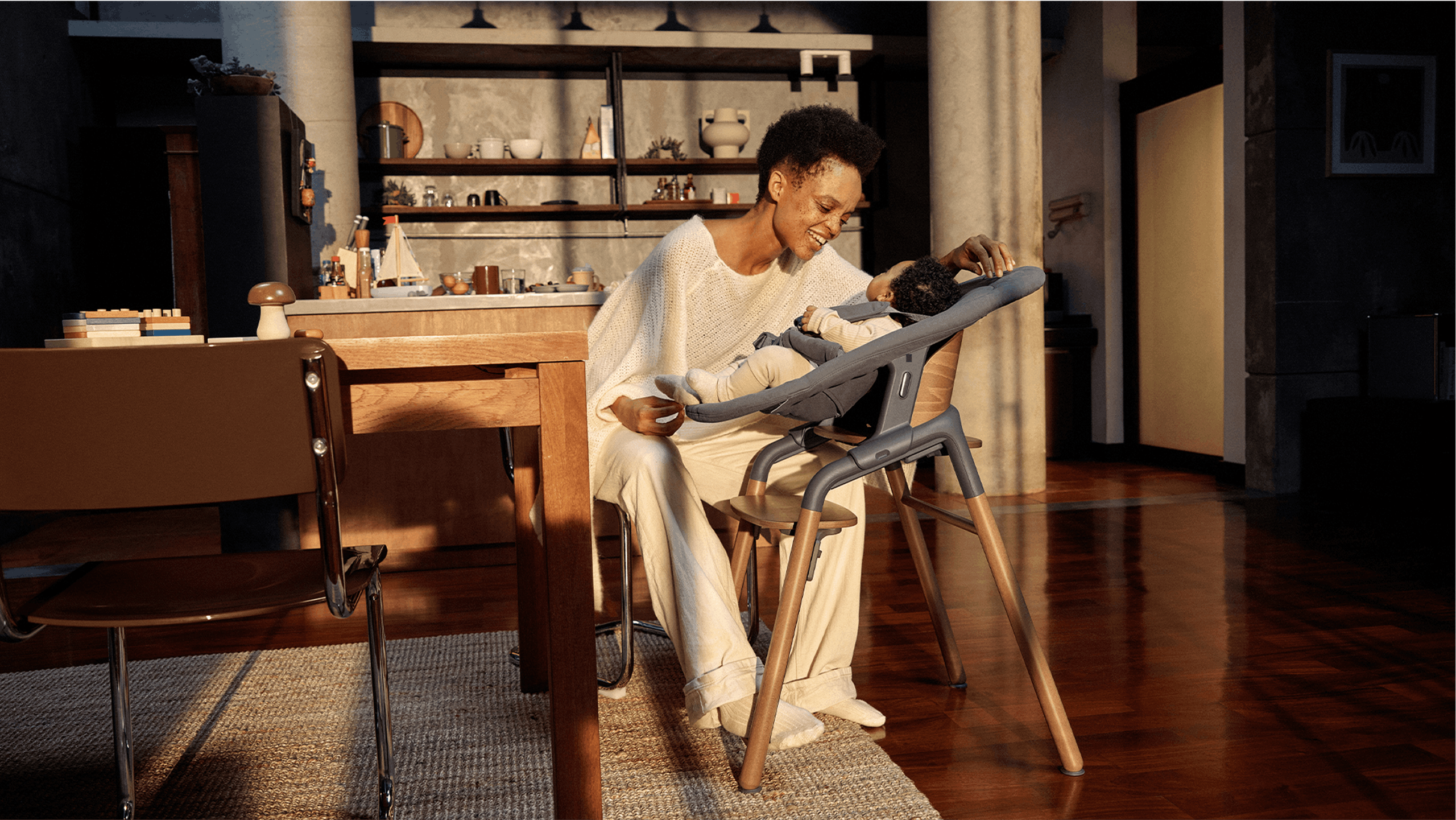 バガブージラフ ハイチェアに座る彼女の赤ちゃんと遊ぶ、カジュアルな服装の母親。 彼らは、暖かみのある照明に包まれたモダンなキッチンにいる。