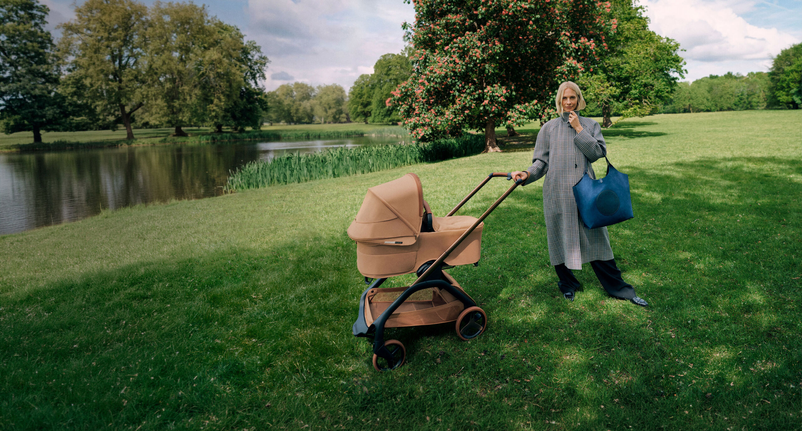 Poppy Delevingne stands next to a Bugaboo x Stella McCartney stroller, holding a large blue tote bag and wearing a long plaid coat. Behind her are a flowering tree, grassy fields, and a pond.