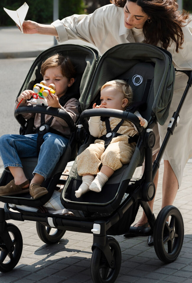 A mother is pushing a Bugaboo Donkey 5 Duo double stroller with her baby and toddler sitting side by side. She leans down while holding a paper airplane to entertain the children.