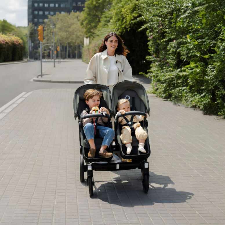 A mom walks along a tree-lined sidewalk pushing a Bugaboo Donkey 5 Duo double stroller with her two children sitting side by side.