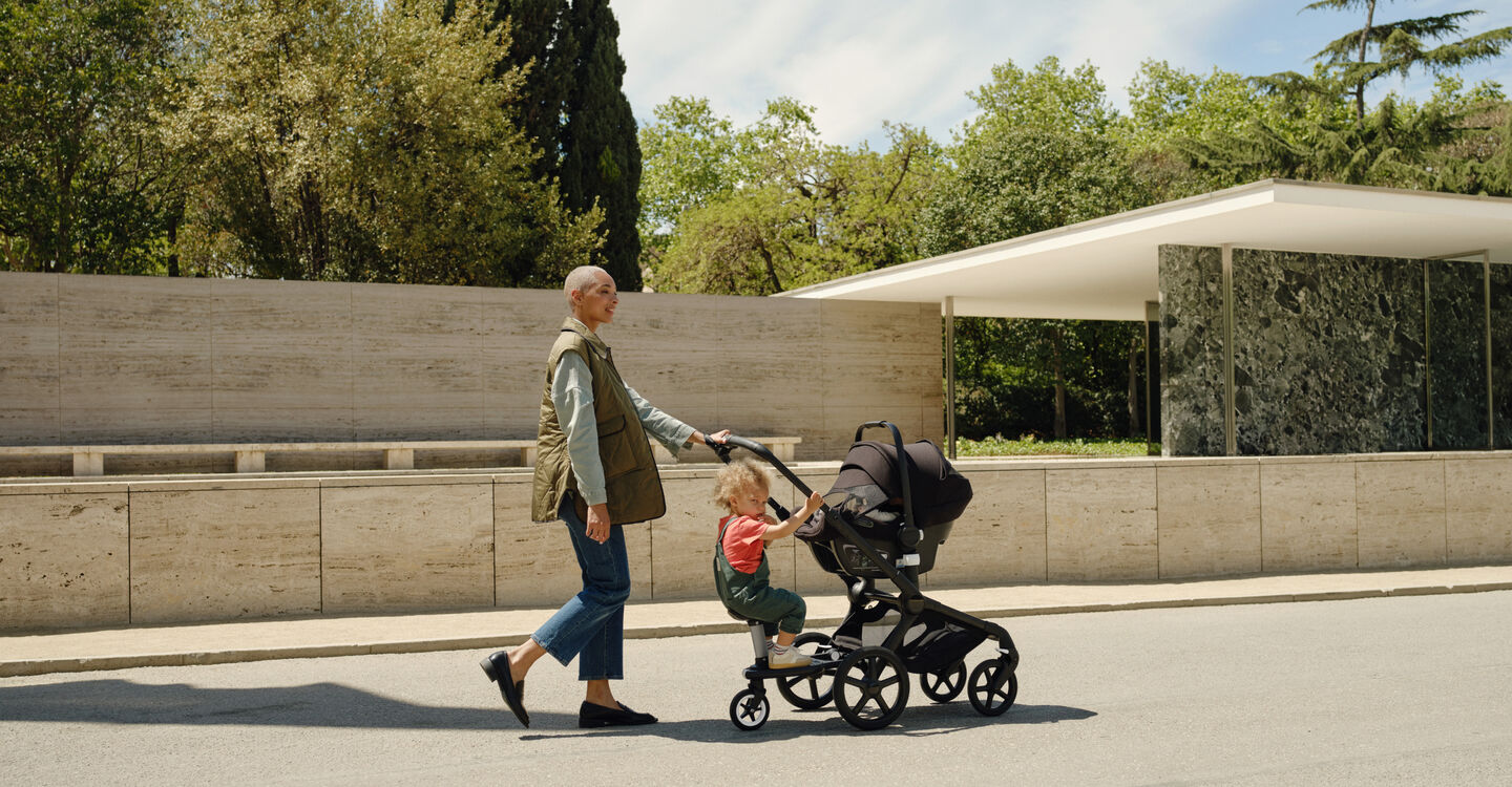 A stylish mother takes a stroll with her kids: Her baby is in a Bugaboo Turtle Air by Nuna attached to a Bugaboo Fox 5, while her toddler rides along on the Bugaboo wheeled board.