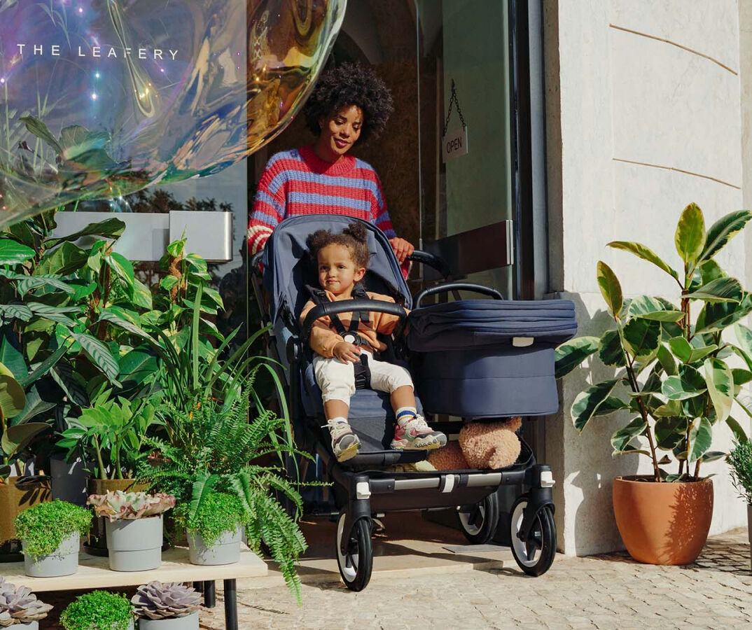 En mamma lämnar en butik och kör en Bugaboo Donkey 5 Duo med en sittdel och en liggdel. Ett barn sitter i sittdelen. Butikens entré är omgiven av grönska.